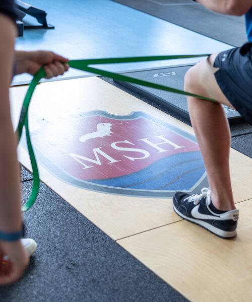 Personen beim Training mit einem Fitnessband in einem Sportraum mit MSH-Logo auf dem Boden.