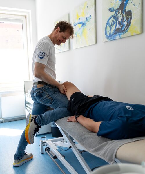 Therapeut behandelt das Bein eines liegenden Patienten in einem hellen Raum mit sportmedizinischen Bildern an der Wand.